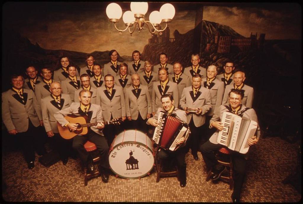 GROUP PHOTOGRAPH OF THE CONCORD SINGERS, MEMBERS OF THE COMMUNITY OF NEW ULM, MINNESOTA, WHO SPECIALIZE IN GERMAN SONGS THE TOWN WAS FOUNDED IN 1854 BY A GERMAN IMMIGRANT LAND COMPANY THE GROUP IS MADE UP OF MEN OF VARYING OCCUPATIONS, AND INCLUDES NEWCOMERS WHO WORK IN THE MANUFACTURING PLANTS AS WELL AS LIFE-LONG RESIDENTS. TWENTY YEARS AGO MANY OF THE TOWN'S INHABITANTS COULD SPEAK SOME GERMAN, BUT THE TRADITION IS DYING AND THE SINGERS ARE TRYING TO KEEP THE HERITAGE ALIVE